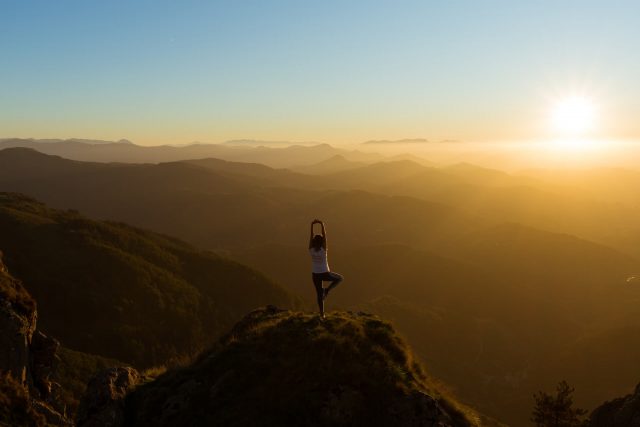 Una persona haciendo una postura de yoga en la cima de una montaña con el amanecer frente a ella, abrazando la serenidad de la naturaleza.